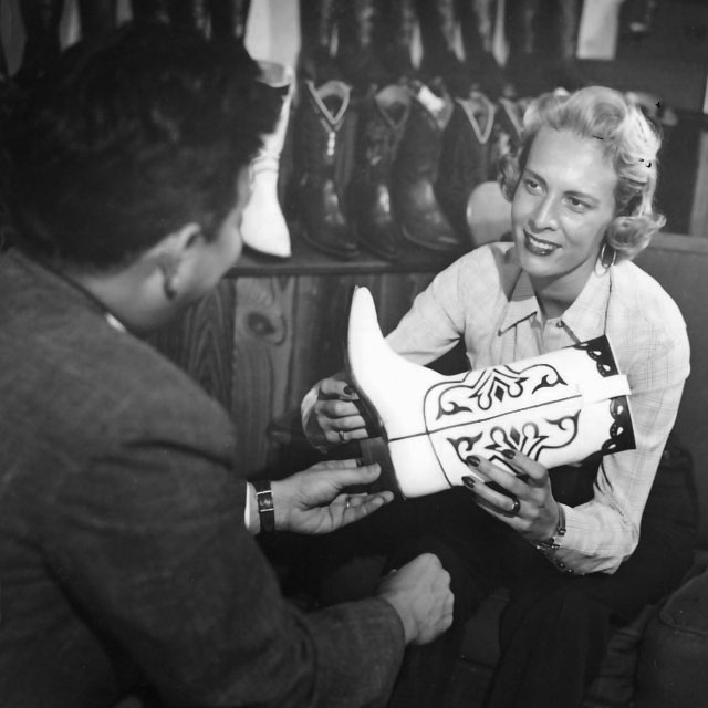 A black and white photo of a young Jane Chilton Justin with blonde hair, wearing business attire and holding a cowboy boot, presenting a boot to a customer.